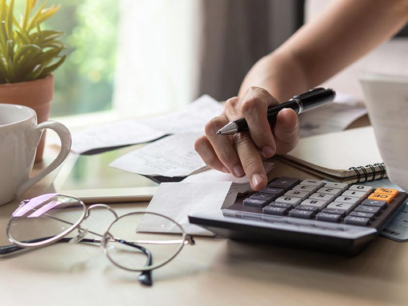 Close view of a hand holding a pen and using a calculator with receipts in the background and glasses and a mug in the foreground.