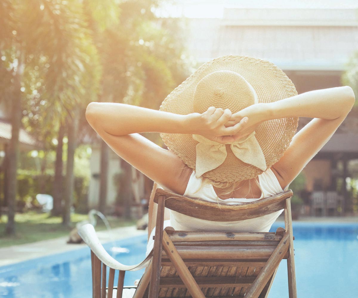 Rear-view of a blonde woman in a straw sun hat, sitting in a wooden deck chair by a pool.